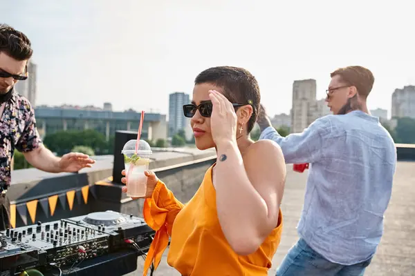 Cheerful beautiful woman with tattoo and sunglasses holding cocktail at party next to DJ equipment — Stock Photo