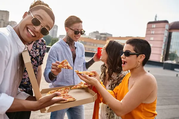 Alegre afroamericano hombre mirando a la cámara mientras disfruta de pizza con sus amigos en la azotea - foto de stock