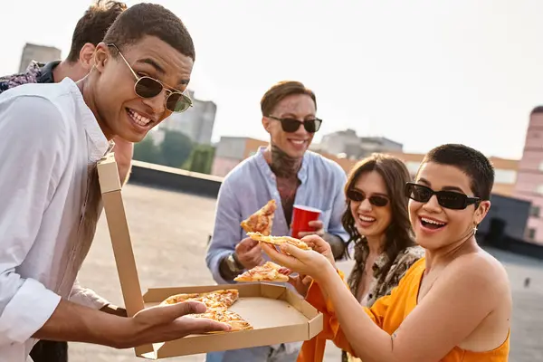 Amigos inter-raciais alegres com óculos de sol sorrindo para a câmera segurando pizza na festa no telhado — Fotografia de Stock
