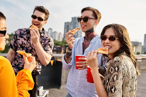 Jóvenes amigos alegres en atuendos vibrantes casuales comiendo pizza y disfrutando de bebidas en la fiesta en la azotea - foto de stock