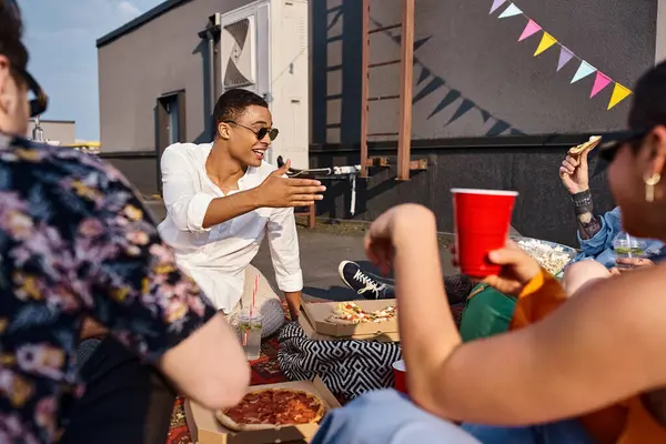 Веселі міжрасові друзі в яскравих вбраннях з сонцезахисними окулярами насолоджуються піцою та напоями на вечірці — стокове фото