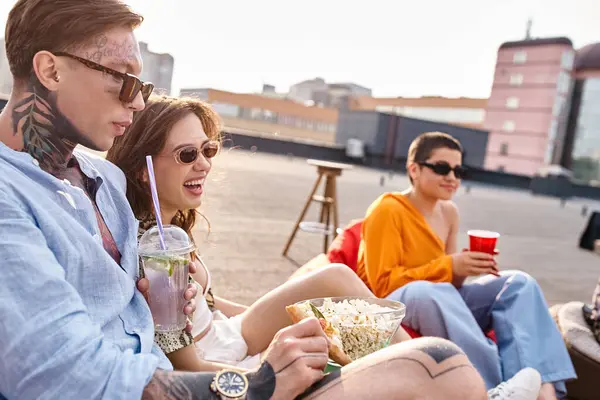Good looking jolly friends in urban attires sitting on rooftop eating popcorn and drinking cocktails — Stock Photo