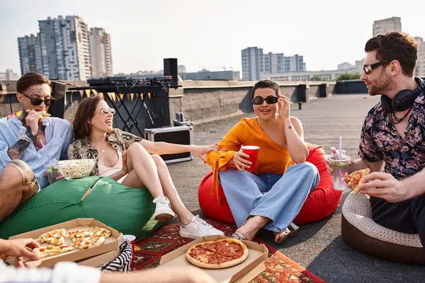 Amigos alegres de buen aspecto en trajes urbanos sentados en la azotea comiendo pizza y bebiendo cócteles - foto de stock