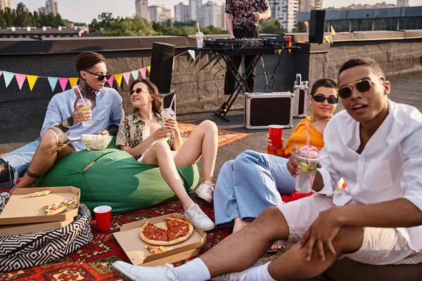 Amigos multiculturales alegres con gafas sentados en la azotea con cócteles y red de pizza para DJ - foto de stock