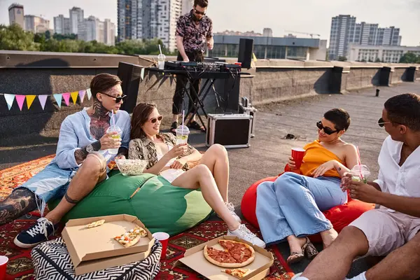 Веселі міжрасові друзі в окулярах сидять на даху з коктейлями та мережею піци для DJ — стокове фото