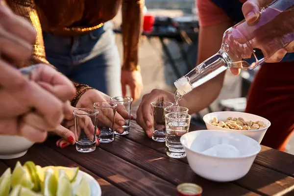 Cropped view of multicultural friends in casual attires pouring tequila shots while at rooftop party — Stock Photo