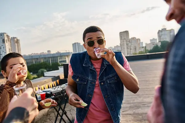 Jolly african american man in vest drinking tequila with lime and salt next to his friends at party — Stock Photo