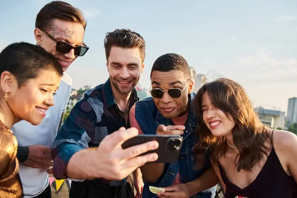 Interracial joyful friends in casual clothes watching photos at smartphone at rooftop party — Stock Photo