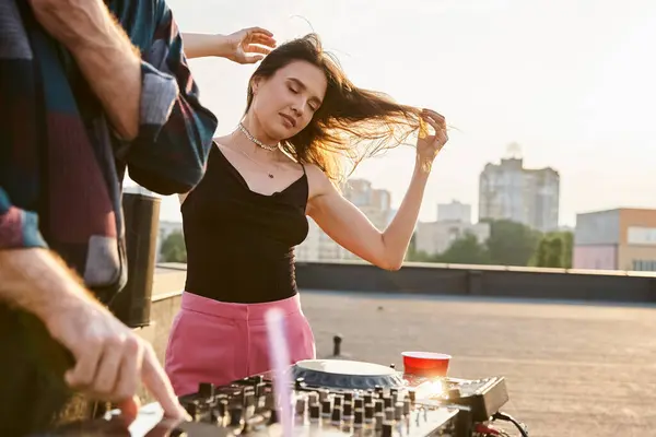 Belle femme aux cheveux longs en tenue vibrante festive posant à côté de DJ pendant la fête sur le toit — Photo de stock