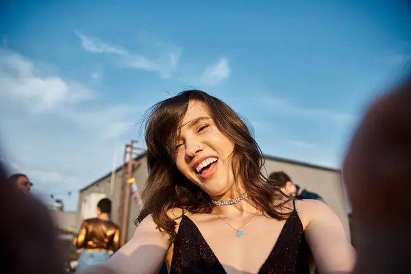 Focus on jolly beautiful woman looking at camera at rooftop party with her diverse blurred friends — Stock Photo