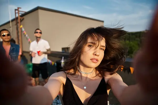 Focus on good looking woman looking at camera during rooftop party with her diverse blurred friends — Stock Photo