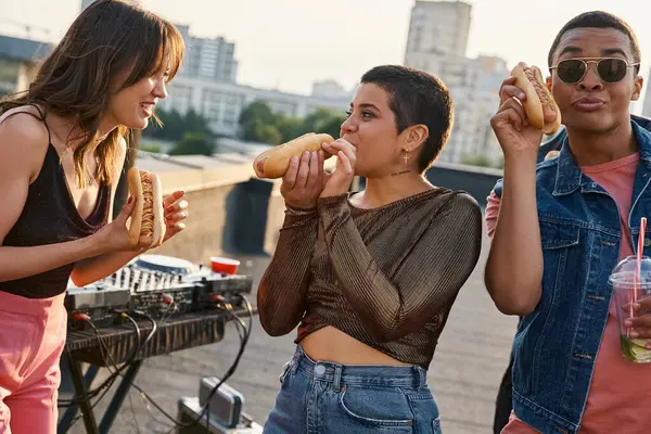 Good looking joyous interracial friends in casual outfits eating tasty hot dogs at rooftop party — Stock Photo
