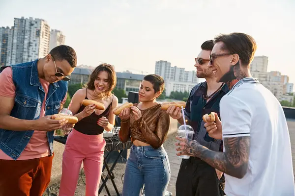Bello cercando amici multiculturali allegri in abiti urbani mangiare gustosi hot dog alla festa sul tetto — Foto stock