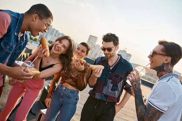 Fröhliche multikulturelle Menschen in lebendigen Outfits genießen köstliche Hot Dogs auf dem Dach bei einer Party — Stockfoto
