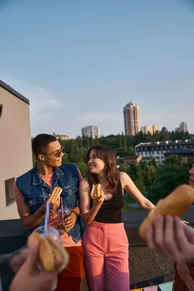 Jolly multiracial friends in vibrant outfits enjoying delicious hot dogs on rooftop at party — Stock Photo