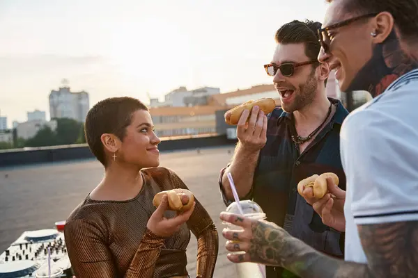 Jolly attractive people with stylish sunglasses in vivid outfits enjoying hot dogs at rooftop party — Stock Photo