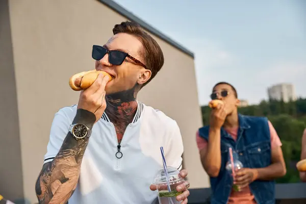 Focus on handsome man with tattoos with his african american friend on backdrop eating hot dogs — Stock Photo