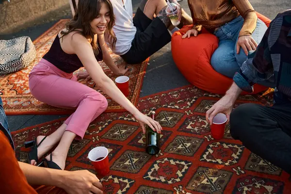Cheerful young multicultural friends in vivid attires playing spin the bottle at rooftop party — Stock Photo