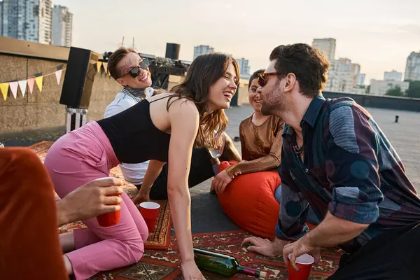 Young jolly people in urban clothes playing spin the bottle together at rooftop, going to kiss — Stock Photo