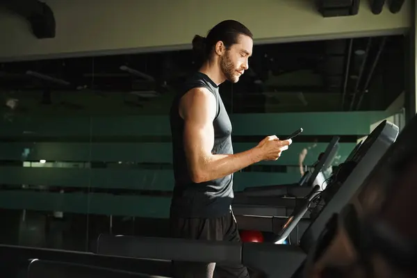 Ein athletischer Mann in aktiver Kleidung steht auf einem Laufband, während er im Fitnessstudio mit seinem Handy trainiert. — Stockfoto