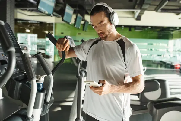 Un hombre enfocado en ropa deportiva que usa auriculares y usa un teléfono celular mientras hace ejercicio en un gimnasio. - foto de stock