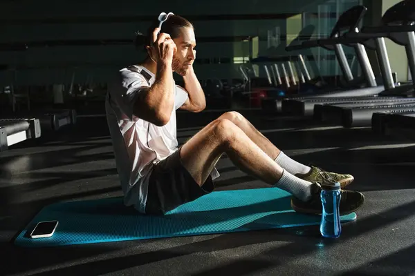 A man in active wear sits on a blue mat, engaged in a music in gym — Stock Photo
