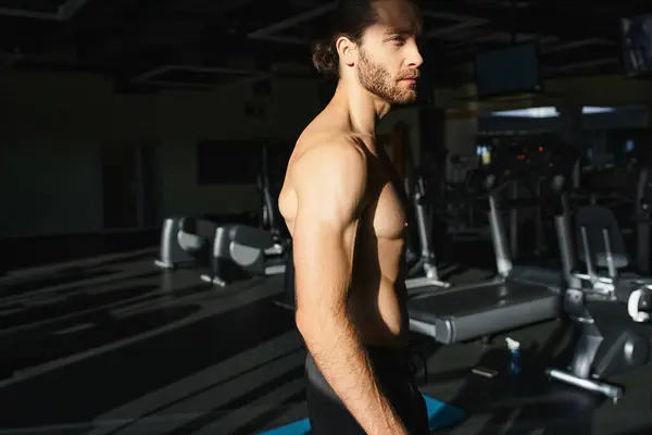 Un hombre musculoso y sin camisa se para con confianza frente a una línea de cintas de correr, listo para una poderosa sesión de entrenamiento.. - foto de stock