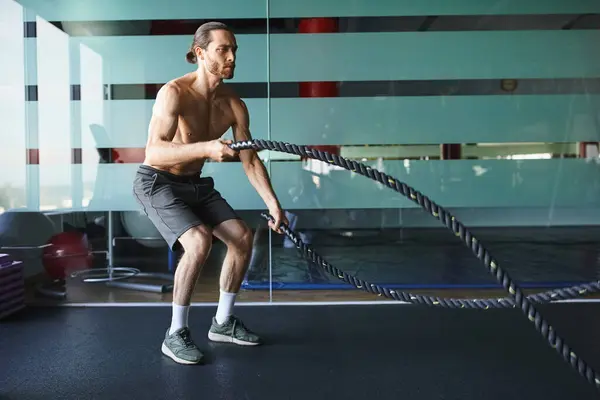 Un hombre musculoso y sin camisa sostiene con confianza una cuerda de batalla en un gimnasio, mostrando su fuerza y determinación. - foto de stock