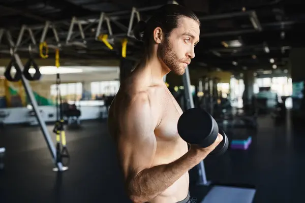 Мужчина без рубашки в спортзале, держащий гантель, демонстрирующий свое мускулистое телосложение и преданность фитнесу. — стоковое фото