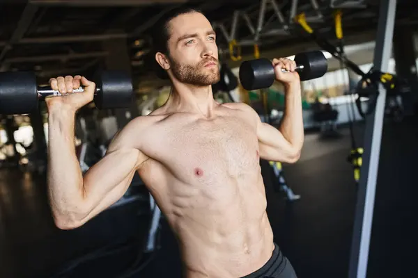 М'язистий чоловік без сорочки тримає двох гантелів у спортзалі, демонструючи свою рутину тренування та фізичну силу. — стокове фото