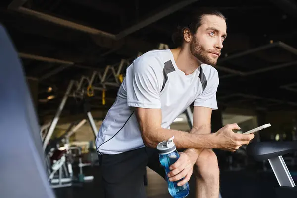 Muskulöser Mann sitzt auf einer Bank, hält eine Flasche Wasser und ein Smartphone in der Hand — Stockfoto