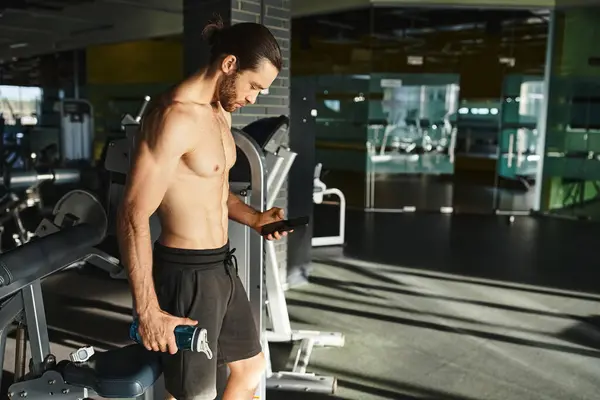 Hombre sin camisa en el gimnasio, intensamente enfocado en la pantalla del teléfono celular mientras hace ejercicio. - foto de stock