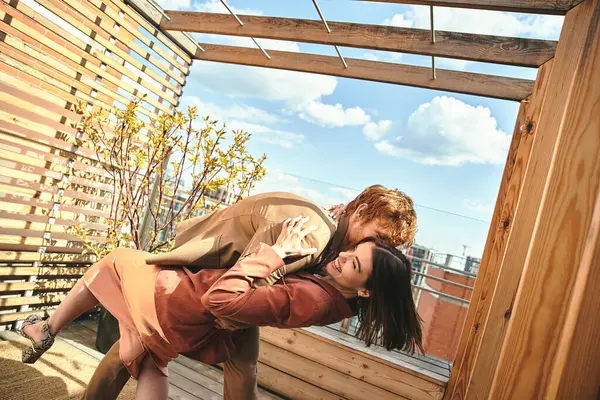 Un homme et une femme s'embrassent paisiblement sur une terrasse en bois surplombant la nature, leurs silhouettes se mélangeant à la lumière du coucher de soleil chaud — Photo de stock