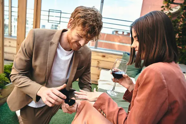 Un hombre arrodillado regalando un anillo a su alegre compañero, compartiendo vino y sonrisas. - foto de stock