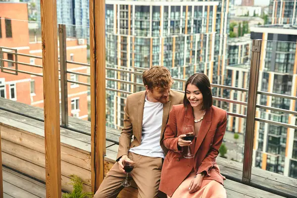 A couple shares a moment with laughter and red wine, framed by cityscape views on a wooden balcony — Stock Photo