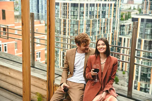 Un couple souriant se détend avec un verre de vin, embrassé par la chaleur du soleil couchant et le paysage urbain — Photo de stock