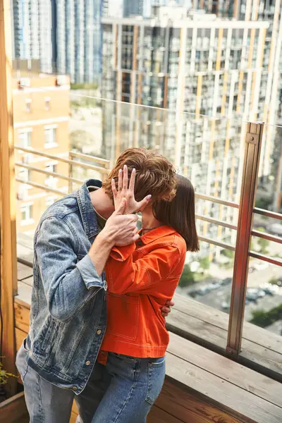 Un hombre y una mujer comparten un tierno beso en un balcón con vistas a un paisaje urbano, sus cuerpos unidos en un abrazo íntimo - foto de stock