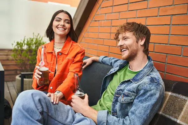 Двоє друзів, сміючись і розмовляючи, насолоджуються освіжаючими напоями на затишному відкритому дивані — стокове фото