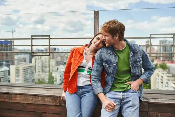 Un hombre y una mujer se sientan juntos en la parte superior de un edificio, con vistas al paisaje urbano mientras comparten un momento de intimidad y conexión - foto de stock