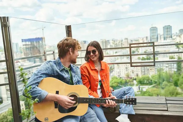 A man with a guitar sings to a smiling woman on a rooftop overlooking the urban skyline. — Stock Photo