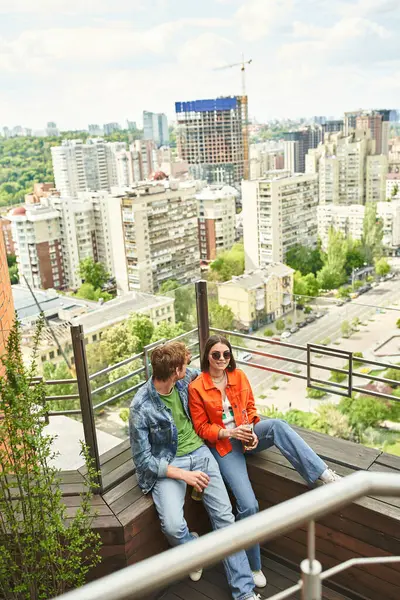 Un par de personas se sientan tranquilamente en el borde de un edificio alto, disfrutando de la vista del horizonte de la ciudad al atardecer. - foto de stock