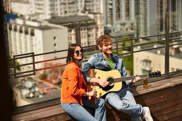 Un hombre y una mujer se sientan en un banco, ella sostiene una guitarra mientras él escucha atentamente. Comparten melodías bajo el cielo abierto - foto de stock
