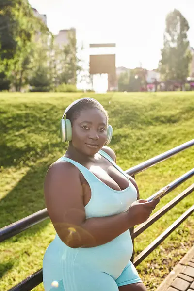 Uma mulher afro-americana com fones de ouvido fica perto de uma cerca, desfrutando de música ao ar livre em um momento positivo, curvilíneo e positivo para o corpo. — Fotografia de Stock