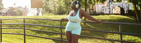 Mulher afro-americana em uma roupa azul, inclinando-se graciosamente em uma cerca, desfrutando de música através de fones de ouvido em um ambiente ao ar livre. — Fotografia de Stock