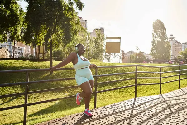 Una mujer afroamericana con auriculares, abrazando la positividad corporal, corriendo en top blanco y pantalones cortos azules. - foto de stock