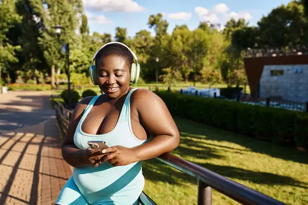 Una mujer afroamericana con auriculares sumergidos en la pantalla de su teléfono celular, disfrutando de la música al aire libre. - foto de stock