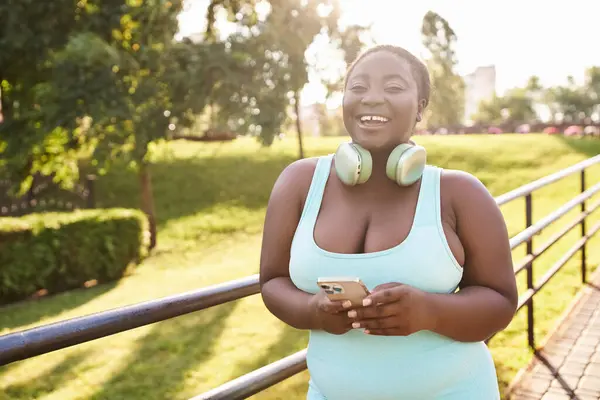 Une femme afro-américaine confiante, portant un casque, aime la musique tout en tenant un téléphone portable à l'extérieur. — Photo de stock