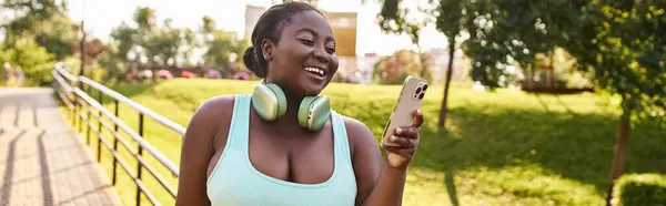 Uma mulher afro-americana ouve alegremente música em seu celular enquanto usa fones de ouvido ao ar livre. — Fotografia de Stock