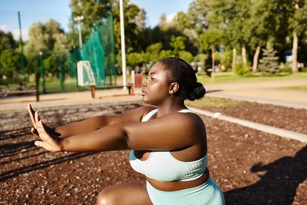 Una mujer afroamericana con un sujetador deportivo azul estira los brazos al aire libre, abrazando su positividad corporal y su viaje de fitness. - foto de stock