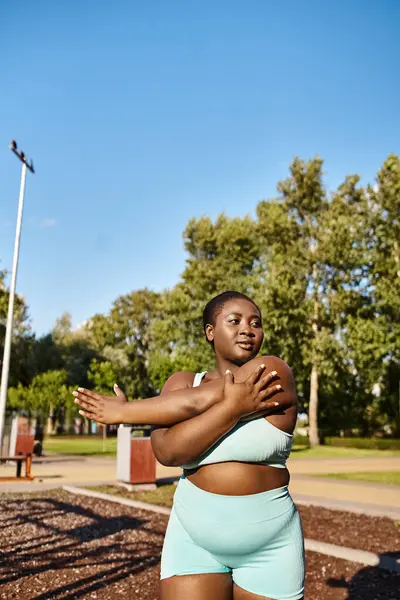 Una mujer afroamericana confiada con un físico curvilíneo se ve usando un sujetador deportivo azul y pantalones cortos, haciendo ejercicio al aire libre. - foto de stock